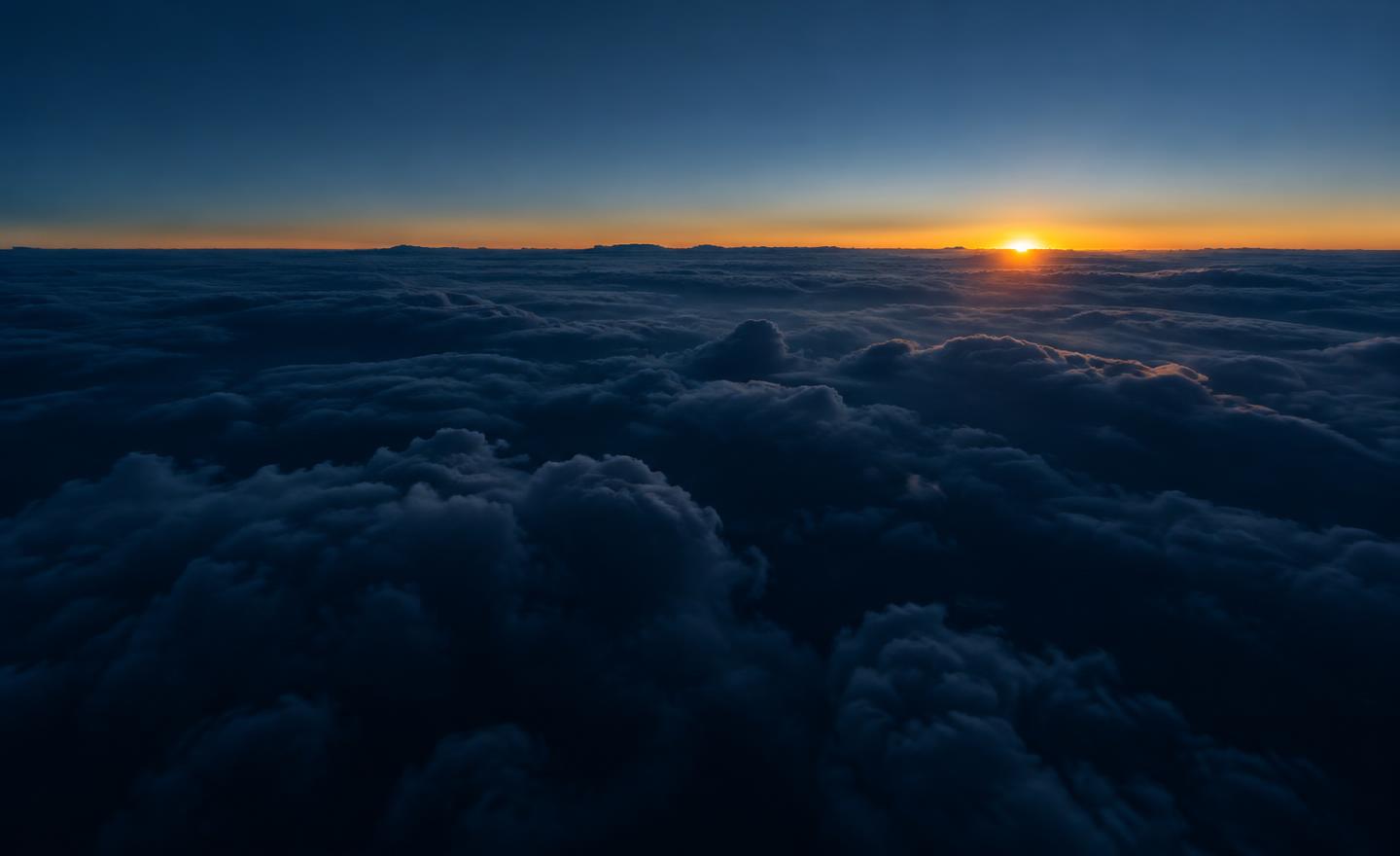 Above a dense cloud layer at dawn, amber light breaking over the horizon